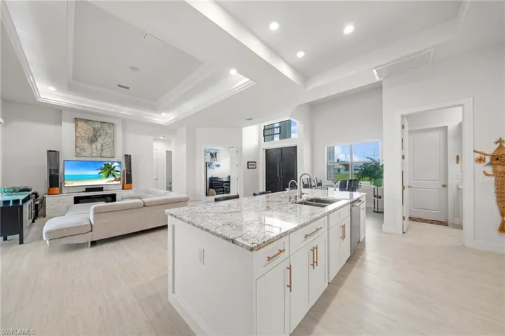 Kitchen featuring a raised ceiling, white cabinetry, light stone counters, an island with sink, and recessed lighting
