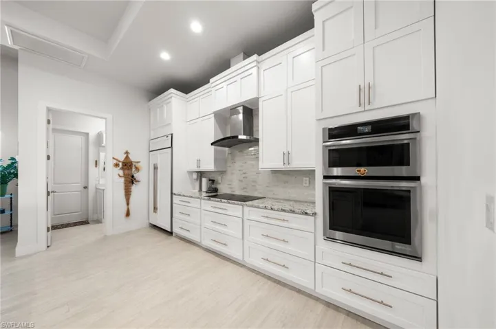 Kitchen featuring stainless steel double oven, decorative backsplash, wall chimney range hood, paneled refrigerator, and light wood finished floors
