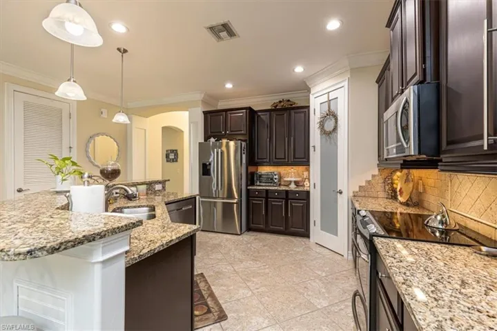 Kitchen featuring crown molding, appliances with stainless steel finishes, dark brown cabinetry, hanging light fixtures, and light stone countertops