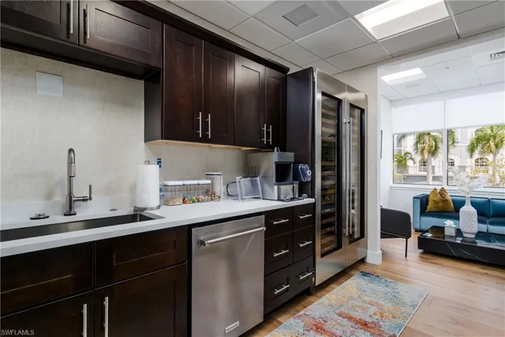 Kitchenette featuring dark wood-finish cabinetry, a stainless steel sink with a gooseneck faucet, light-toned countertops, and a built-in stainless steel dishwasher