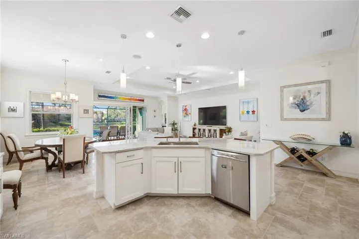 Kitchen featuring stainless steel dishwasher, ceiling fan with notable chandelier, white cabinetry, hanging light fixtures, and an island with sink