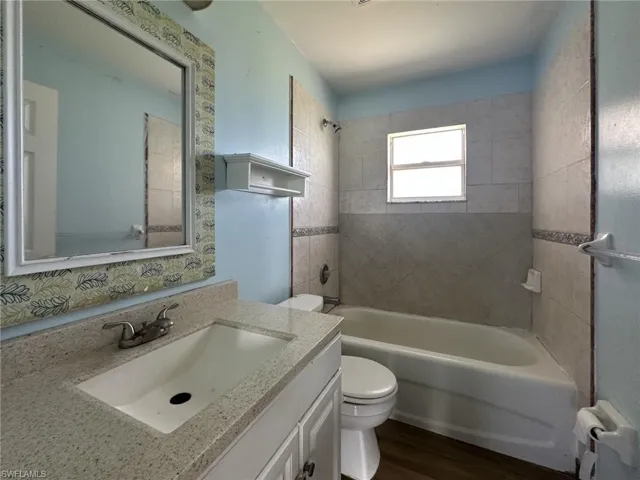 Bathroom featuring a single vanity with an integrated sink and light-toned countertop, a framed wall mirror, a toilet, a bathtub with surrounding tile, and a window providing natural light