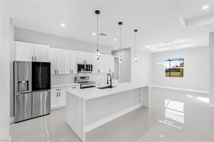 Kitchen with decorative light fixtures, appliances with stainless steel finishes, light stone counters, white cabinets, and an island with sink