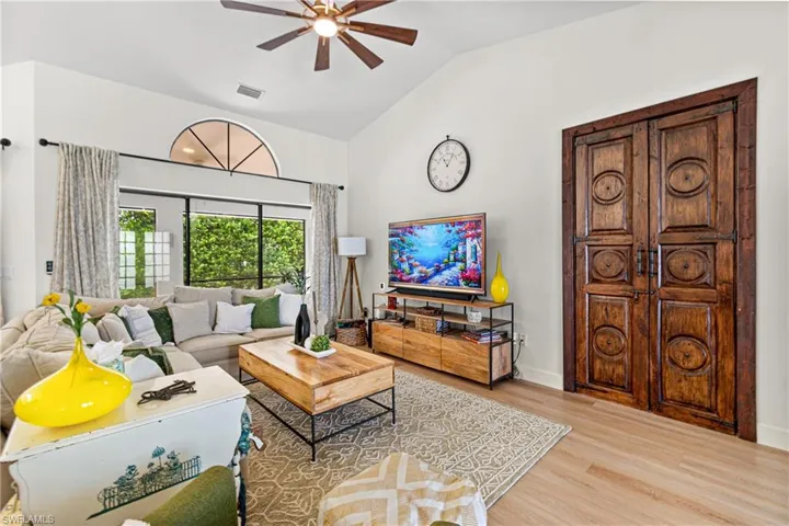 Vaulted ceiling living area featuring a decorative wood-finish ceiling fan, expansive windows with an arched transom, light wood-finish flooring, and ornate double doors