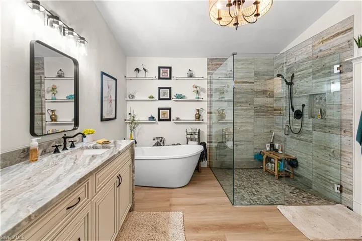 Spacious bathroom featuring a frameless glass shower enclosure with pebble flooring and wood-look tile surround, a freestanding soaking tub, a double vanity with stone countertop and integrated sinks, and wood-finish flooring