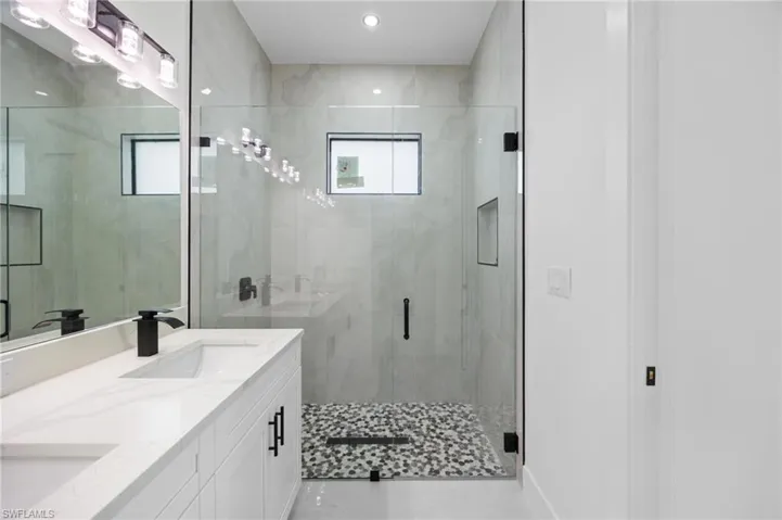 Spacious bathroom featuring a frameless glass shower enclosure, mosaic pebble floor, tiled shower walls, dual vanity with quartz countertop, and black hardware accents