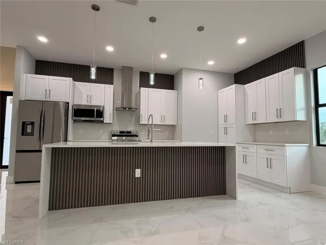 Kitchen featuring a large island with sink, stainless steel appliances, wall chimney exhaust hood, white cabinetry, and hanging light fixtures