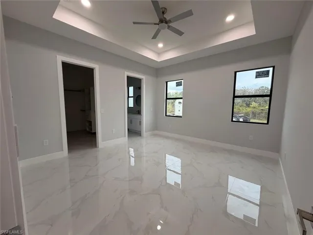 Unfurnished bedroom with a spacious closet, a raised ceiling, light marble finish floors, recessed lighting, and a ceiling fan