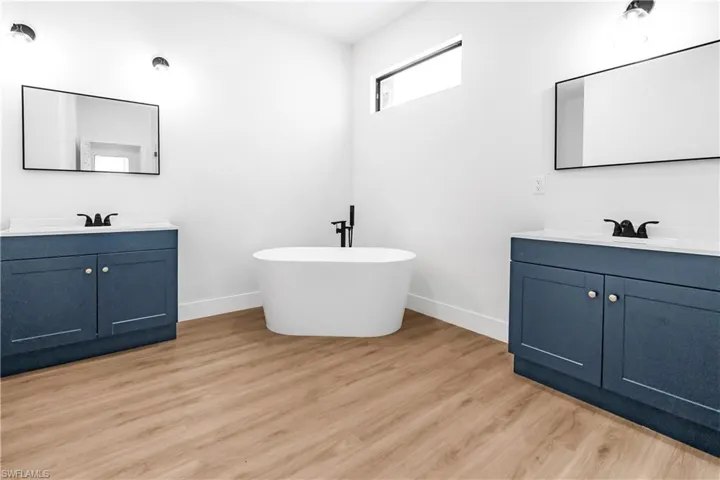 Freestanding soaking tub with matte black fixtures, dual vanity sinks with blue cabinetry, white countertops, black-framed mirrors, and wood-finish flooring