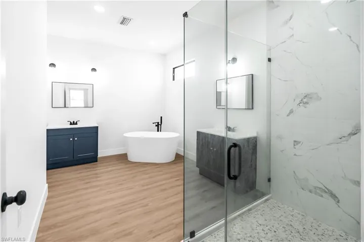 Spacious bathroom featuring light wood-finish flooring, a freestanding soaking tub with a matte black fixture, and a glass-enclosed shower with large format marble-look tiling