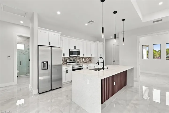 White shaker cabinetry with black hardware, stainless steel appliances, patterned tile backsplash, quartz countertop island with wood-finish paneling, and pendant lighting