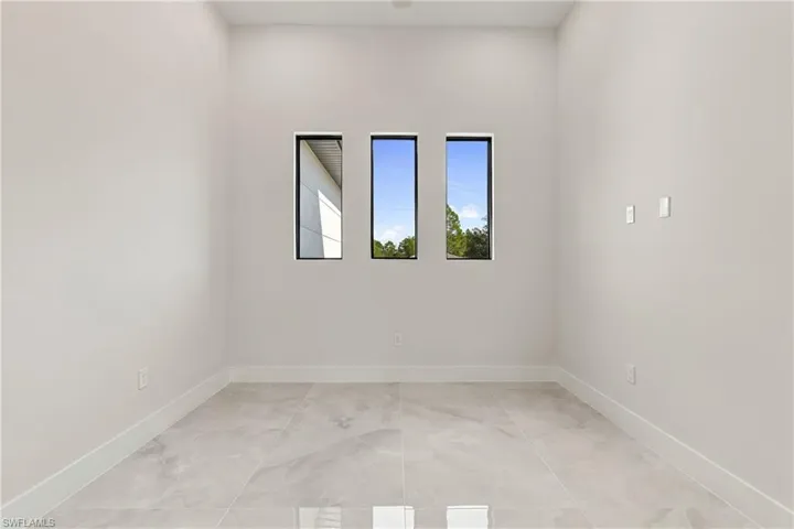 Recessed lighting illuminates a room featuring three vertical windows with dark frames, light-toned walls, and large format floor tiling
