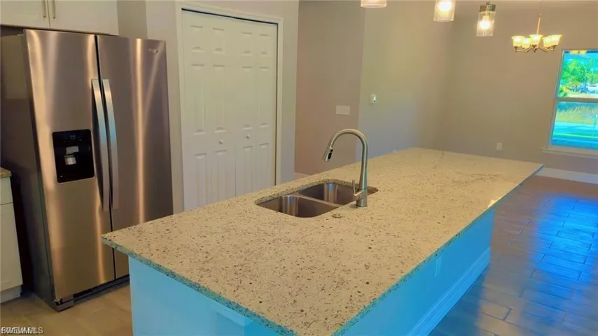 Kitchen featuring stainless steel refrigerator with ice dispenser, light stone counters, a kitchen island with sink, white cabinets, and a chandelier