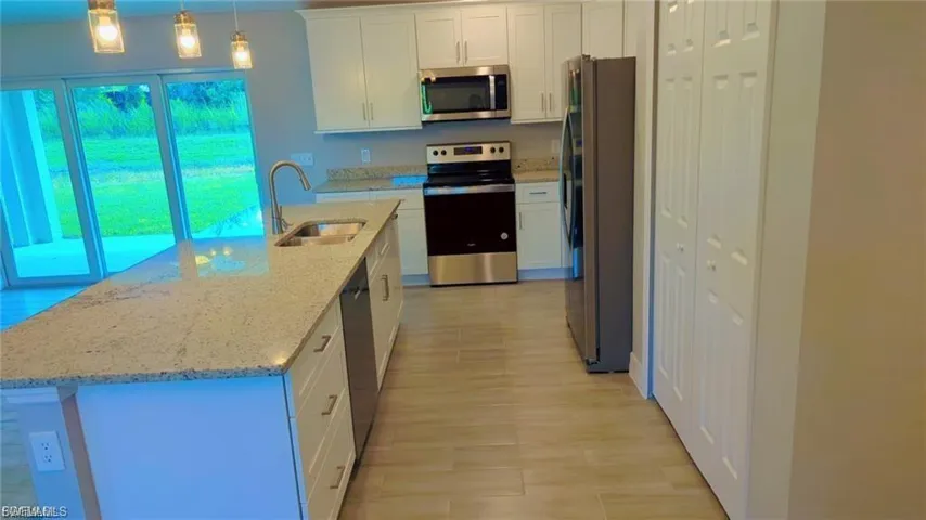 Kitchen featuring white cabinets, stainless steel appliances, a center island with sink, light stone counters, and decorative light fixtures
