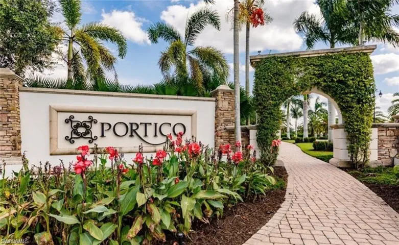Community entrance featuring a stone-clad sign with decorative ironwork, a brick paver pathway, a vine-covered archway, and lush tropical landscaping with palm trees and vibrant red flora