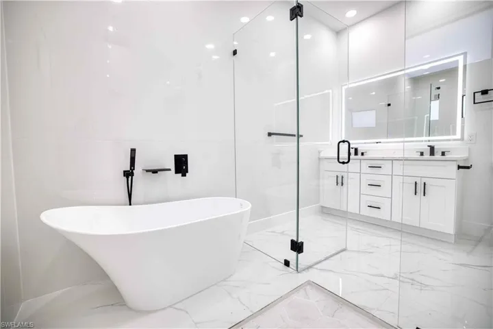 Freestanding soaking tub with wall-mounted matte black faucet, glass-enclosed shower, dual vanity with shaker-style cabinetry, quartz countertop, and illuminated mirror