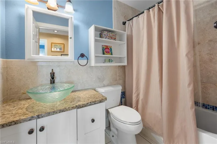 Bathroom featuring a glass vessel sink, granite-style countertop, white vanity cabinetry, wall-mounted storage shelves, and tiled shower surround with accent border
