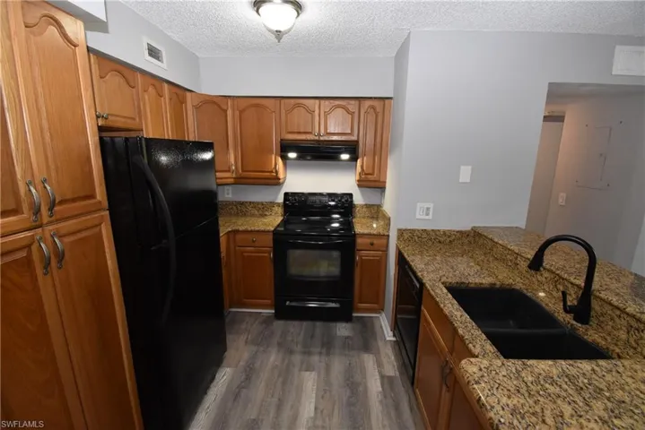 Kitchen with black appliances, wood finish cabinets, dark stone countertops, a textured ceiling, and dark wood-style flooring