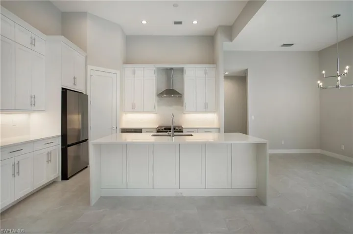 Kitchen with a center island with sink, stainless steel fridge, wall chimney range hood, pendant lighting, and white cabinets