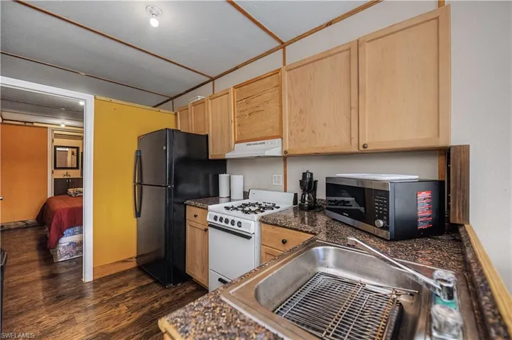 Kitchen featuring light brown cabinetry, white range with gas cooktop, stainless steel microwave, dark wood finished floors, and freestanding refrigerator