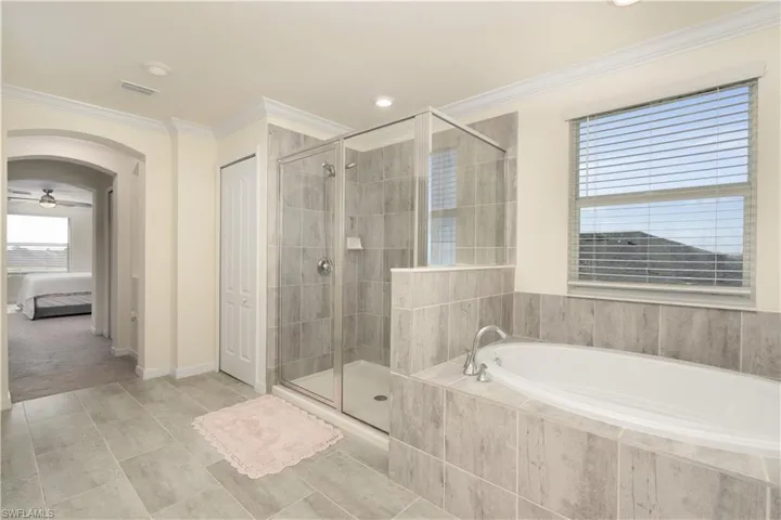 Bathroom featuring a built-in tub with tile surround, a glass-enclosed shower, large window with blinds, tile flooring, and an arched entryway
