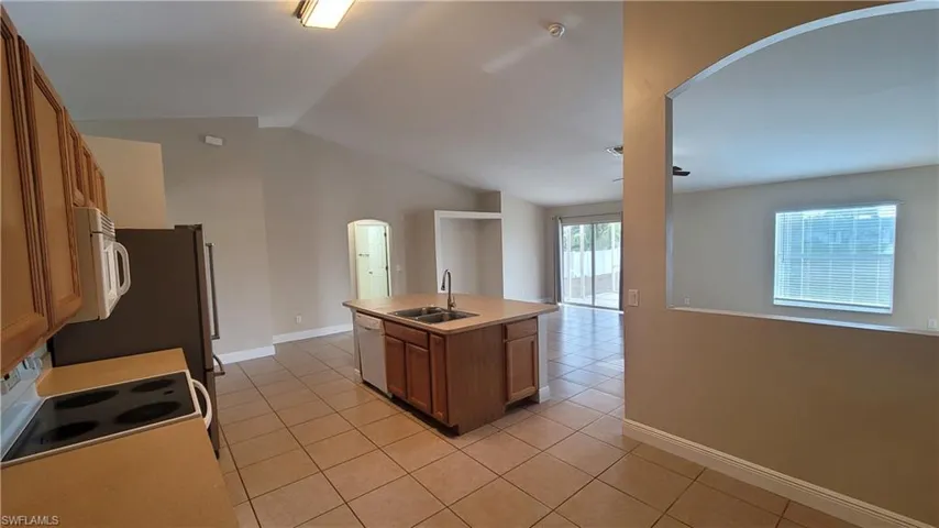 Kitchen featuring arched walkways, wood finish cabinetry, a center island with sink, white appliances, and lofted ceiling