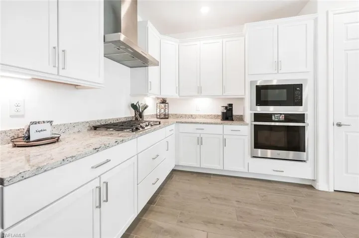 Kitchen with wall exhaust hood, appliances with stainless steel finishes, light stone counters, light tile flooring, and white cabinets