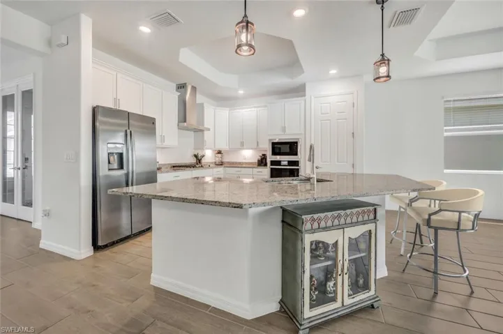 Kitchen with white cabinetry, stainless steel appliances, an island with sink, a tray ceiling, and wall range hood