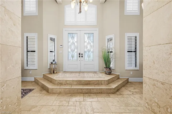 Grand entryway featuring double doors with decorative glass inserts, plantation shutters, a tiered landing, and extensive stone tile flooring
