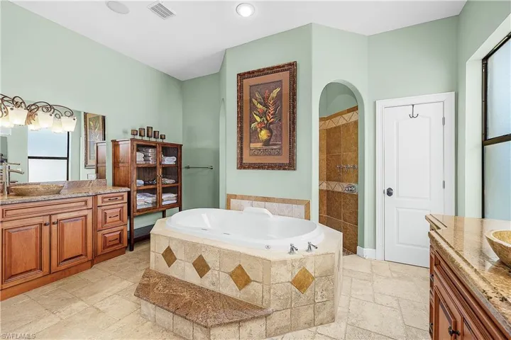 Spacious bathroom featuring a built-in jetted tub with tiled surround, a separate arched shower enclosure, dual wood-finish vanity cabinets with stone countertops, and a light green wall finish
