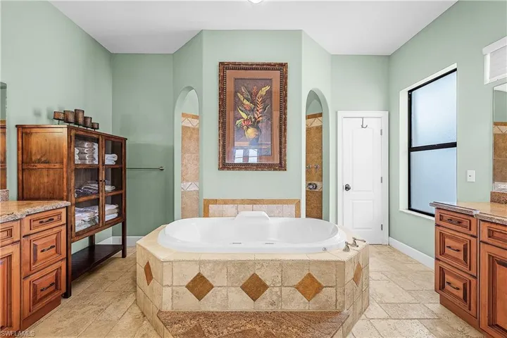 Soaking tub with a tiled surround, arched doorways, and dual vanity areas with wood cabinetry and stone countertops