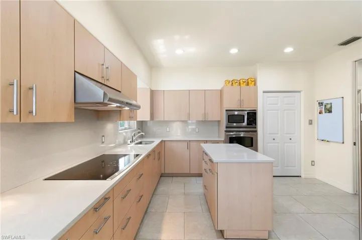 Modern kitchen featuring light wood-finish cabinetry, a central island with a solid surface countertop, a built-in cooktop, and stainless steel appliances