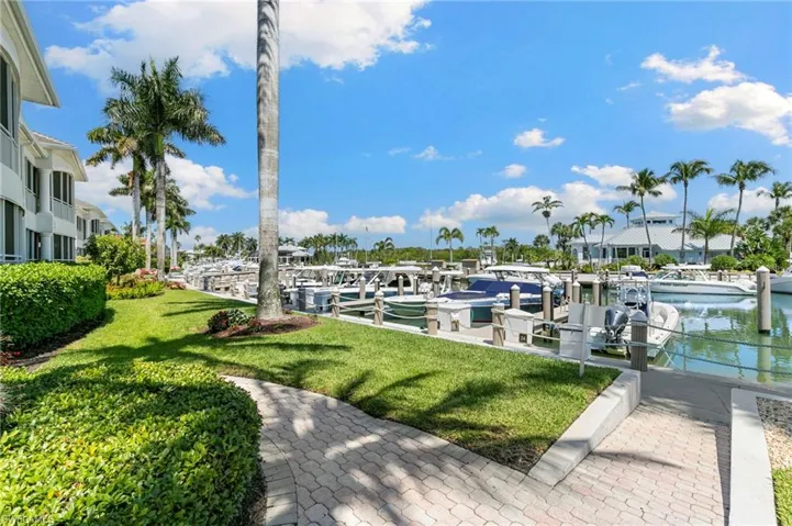Marina featuring multiple boat slips, palm trees, waterfront buildings, green lawn, and a paver pathway