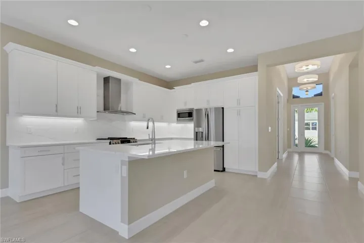 Kitchen featuring backsplash, white cabinets, wall chimney range hood, an island with sink, and light stone countertops