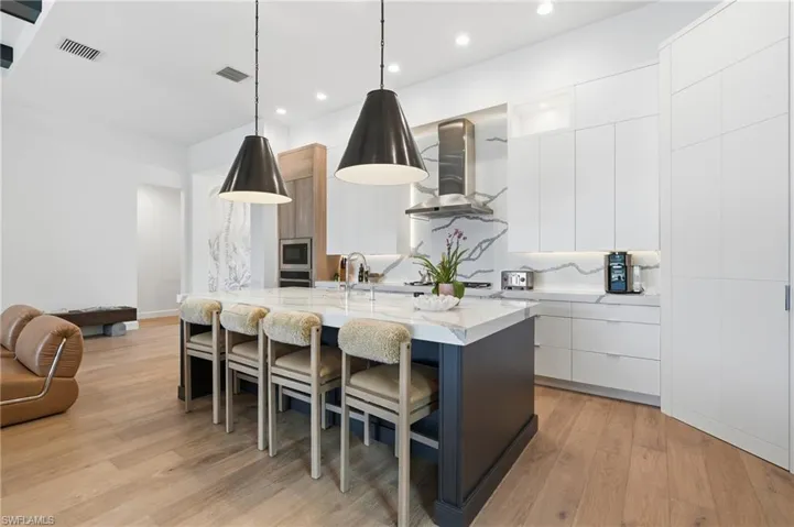 Kitchen featuring a large central island with seating, pendant lighting, light wood-finish flooring, white flat-panel cabinetry, and a full-height backsplash with integrated LED lighting