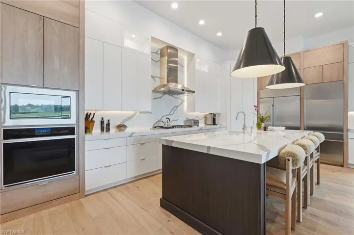 Gourmet kitchen featuring a large central island with seating, integrated stainless steel appliances, extensive white cabinetry, quartz countertops with a matching backsplash, and light wood-finish flooring