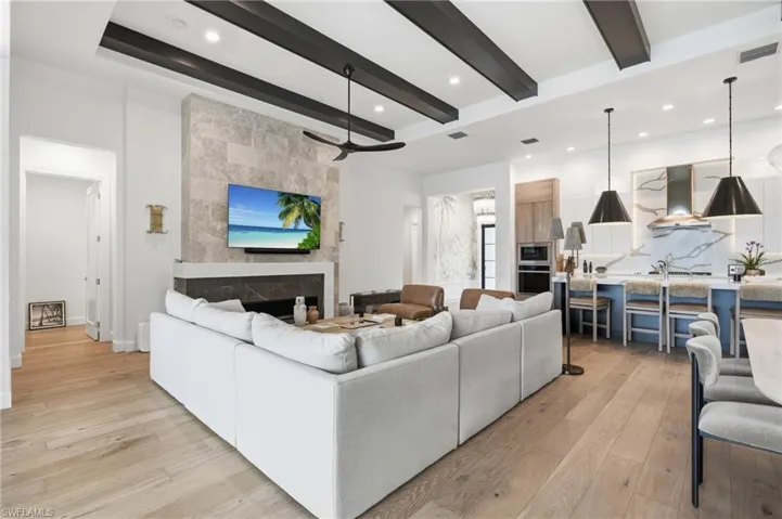 Open-concept living space featuring wood-finish flooring, a contemporary fireplace with a stone surround, exposed ceiling beams, and recessed lighting
