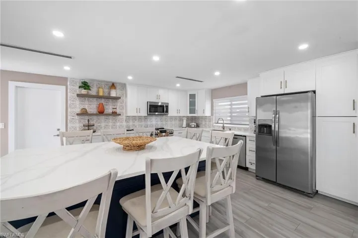 Kitchen featuring white shaker-style cabinetry, patterned tile backsplash, floating wood shelving, quartz-topped center island, and wood-finish flooring