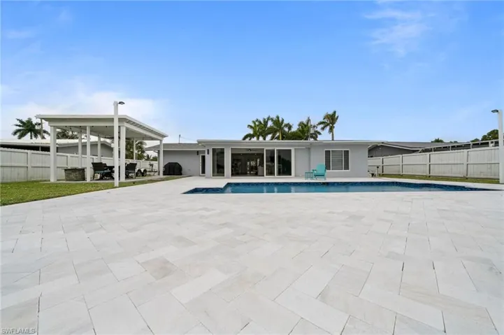 Rear exterior featuring a swimming pool, expansive herringbone-patterned stone patio, covered pergola, and perimeter privacy fencing