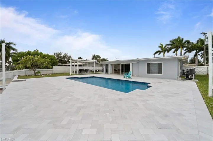 Rear exterior featuring a rectangular in-ground swimming pool, extensive stone paver patio, covered pergola, and white privacy fencing