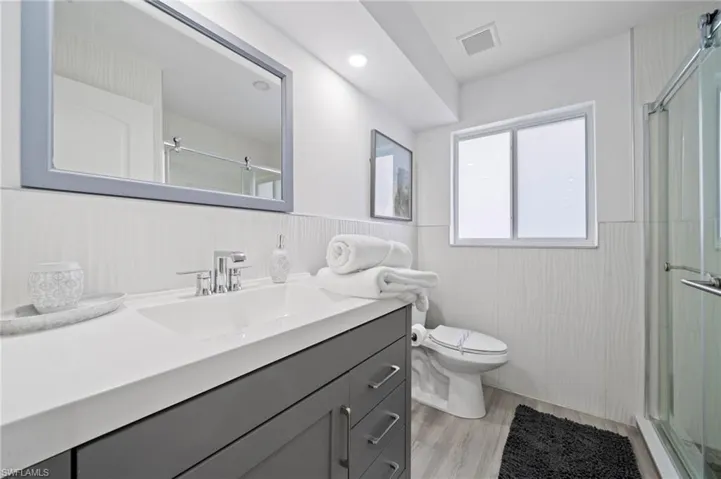 Bathroom featuring a gray vanity with a white countertop and integrated sink, a framed wall mirror, a glass-enclosed shower, and wood-finish flooring