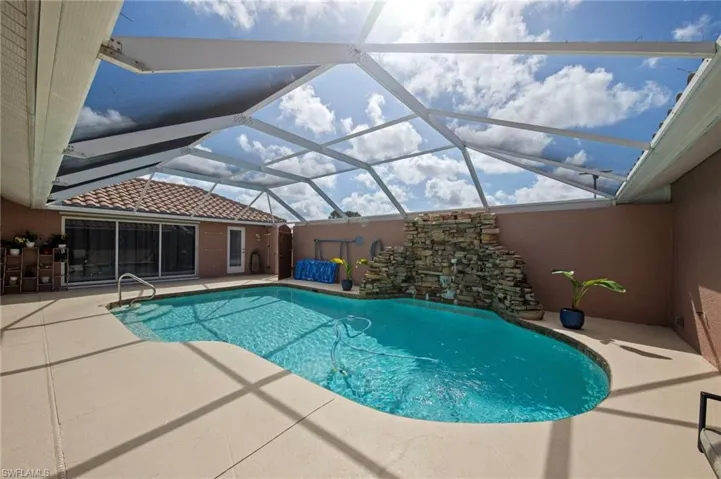 Screened outdoor pool area featuring a freeform swimming pool with integrated waterfall feature, surrounding concrete decking, and an enclosed living space with sliding glass doors