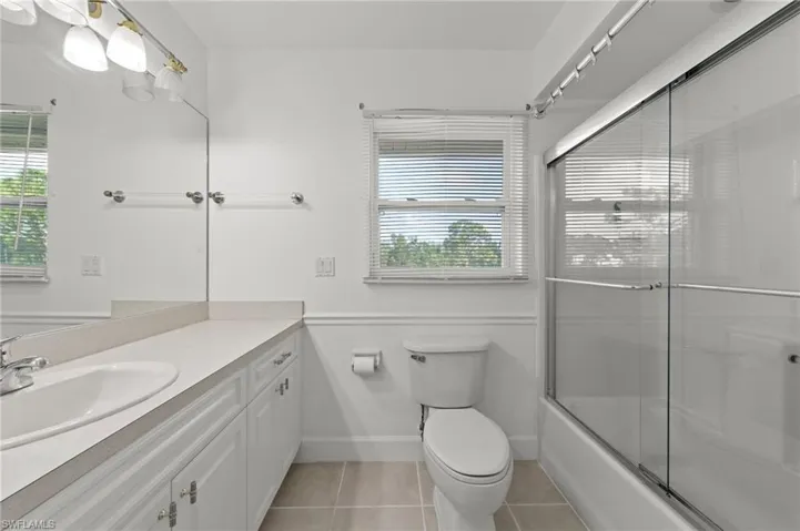 Bathroom featuring a white vanity with an integrated sink, a large wall-mounted mirror, a toilet, a shower-tub combination with sliding glass doors, and two windows with blinds