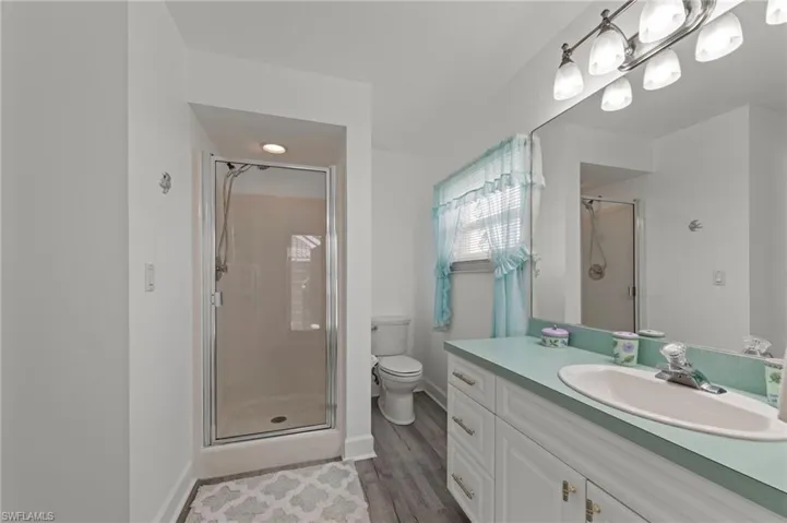 Shower enclosure with clear glass door, single vanity with light teal countertop and integrated sink, white cabinetry with silver-tone hardware, and wood-finish flooring