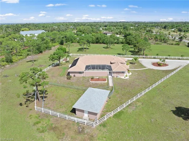 Expansive property featuring a single-story residence with a tan exterior and tile roof, an attached screened enclosure, a detached auxiliary building with a pitched roof, a circular driveway with a landscaped island, and a perimeter white fence enclosing a substantial grassy area