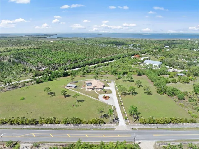 Expansive property with a long, paved driveway leading to a single-story residence, featuring a white fence boundary, mature trees, and a distant water backdrop