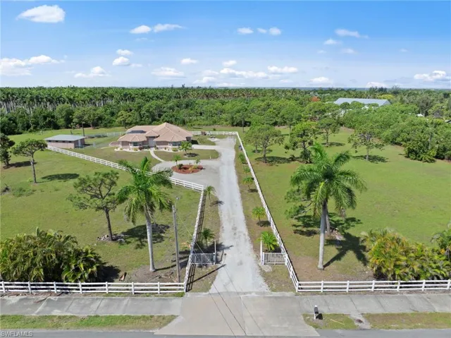 Expansive property featuring a long gravel driveway, white fencing, mature palm trees, and a detached outbuilding
