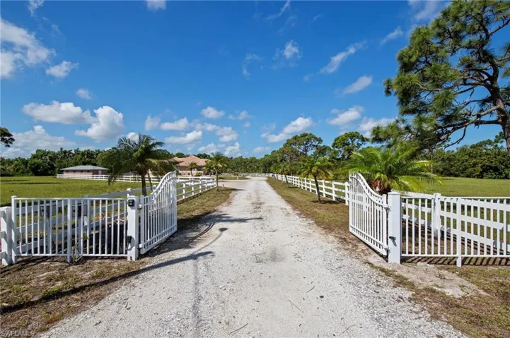 Gated entry with white fencing and a gravel driveway, framed by mature trees and expansive green space