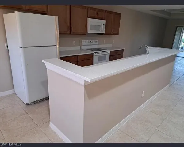 Kitchen with white appliances, light tile patterned flooring, a center island with sink, and brown cabinetry