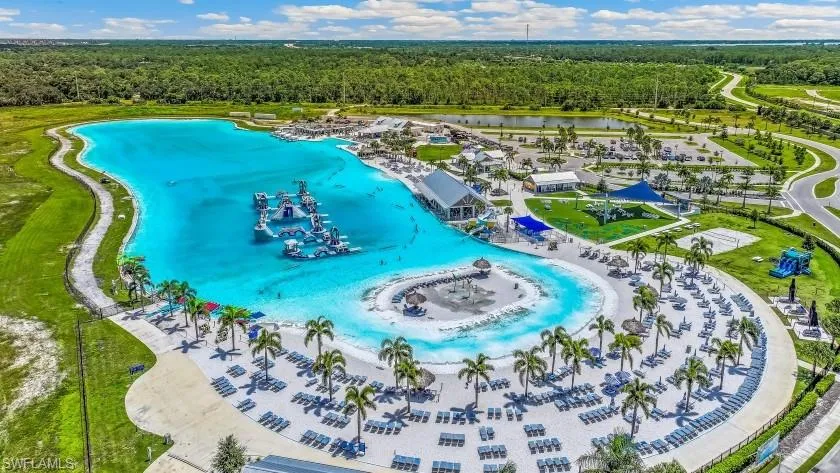 Expansive turquoise lagoon with integrated water features, bordered by a white sand beach with numerous lounge chairs, mature palm trees, and a shaded outdoor structure